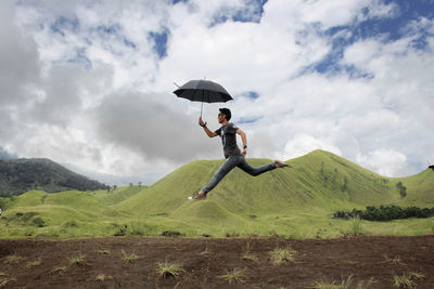 Side view of young man with umbrella jumping by on mountain against cloudy sky