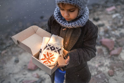 Pretty girl in knitted grey hat opening a crafted gift box with a new pair of gloves, snowfall