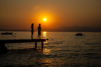 Silhouette men standing in sea against orange sky