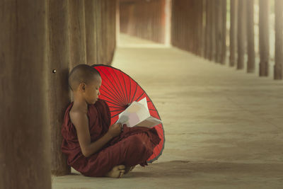 Full length of monk reading book while sitting at corridor