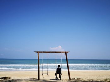 Rear view of teenage boy sitting on swing at beach against blue sky