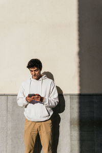 Young smiling male standing on concrete wall in the city street and browsing mobile phone