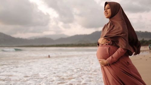 Smiling pregnant woman with hands on stomach wearing hijab standing at beach against cloudy sky