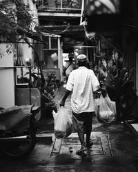 Rear view of man holding plastic bags while walking in alley