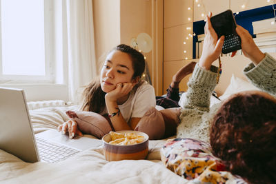 Young woman using laptop while friend text messaging through smart phone on bed