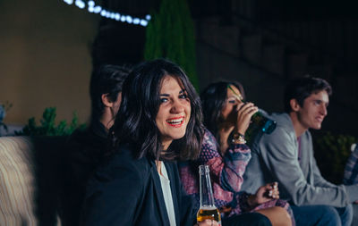 Portrait of cheerful woman having beer with friends at patio during night