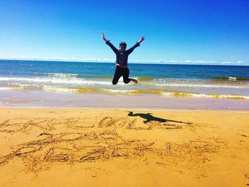 Full length of happy woman jumping over text at beach against clear blue sky