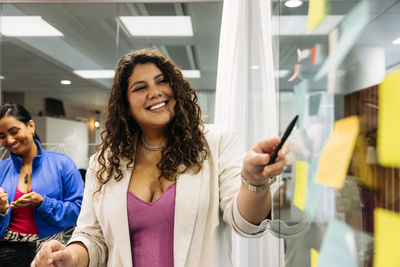 Smiling female business professional pointing at glass wall while doing brainstorming with colleague at office