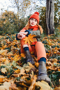 Bright cute young woman in orange warm knitted hat and scarf on background of yellow autumn leaves. 