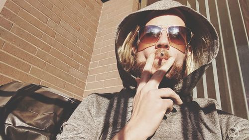 Portrait of man wearing sunglasses and hooded shirt smoking cigarette at home