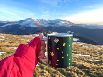 Cropped hand of woman holding mug against mountains