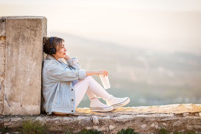 Woman sitting on retaining wall against sky