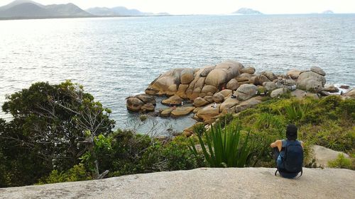 Rear view of man sitting on rock by sea against sky