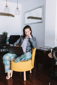 Full body of young female remote employee wearing formal blazer and homey pajama pants sitting on chair and working on laptop at home