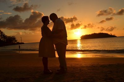 Full length of couple embracing while standing at beach against sky during sunset