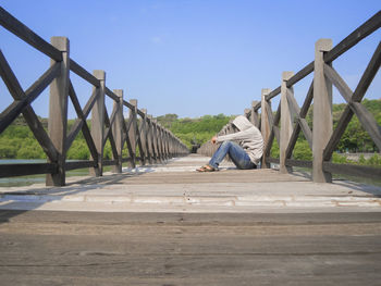 Side view of sad man sitting on footbridge against sky