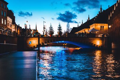 Bridge over river by illuminated buildings against sky at dusk