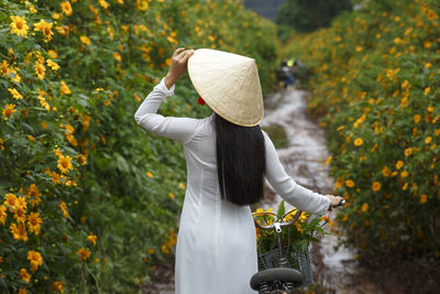 Rear view of woman in asian style conical hat with bicycle amidst flowering plants on field