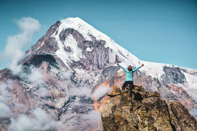 Rear view of woman standing on rock formation with arms outstretched against mountain and sky