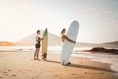 Man and woman standing with surfboard on beach against sea and sky