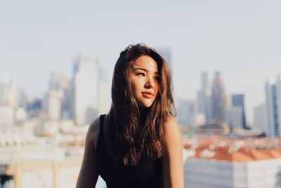 Young woman with long hair looking away while sitting against modern buildings in city