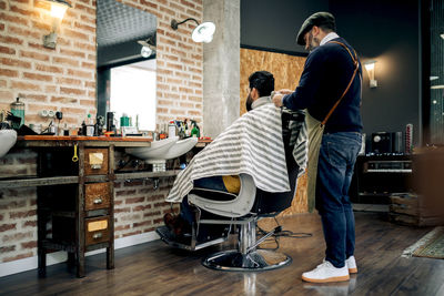 Back view male barber with comb brushing hair of unrecognizable client while making hairstyle in modern barbershop