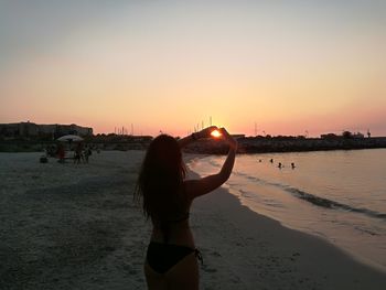 Woman making heart shape with hands at beach against clear sky during sunset