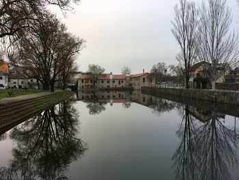 Reflection of bare trees and buildings in lake against sky