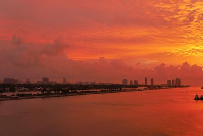 Distant view of city by sea against cloudy sky during sunset