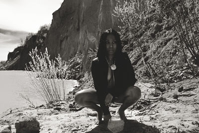 Full length of seductive woman looking away while crouching by lake during sunny day