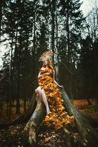 Young woman covered with leaves sitting on tree at forest during autumn