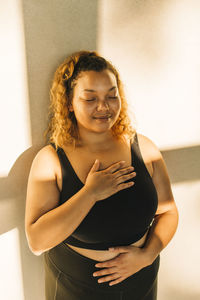 Smiling woman practicing breathing exercise standing against wall