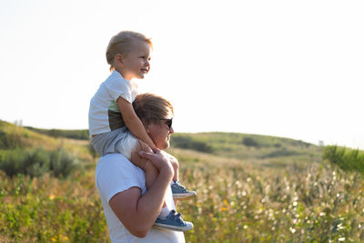 Smiling kid blond boy sitting on dad shoulders in nature on a windy and sunny day