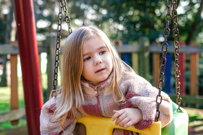 A young girl sits on a swing in a playground, her expression is thoughtful and introspective. 