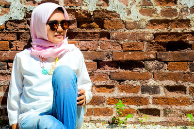 Young woman wearing sunglasses and scarf sitting against brick wall on sunny day
