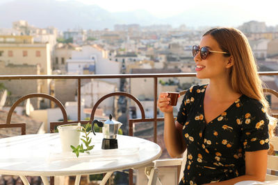 Young woman wearing sunglasses with coffee cup on table against cityscape