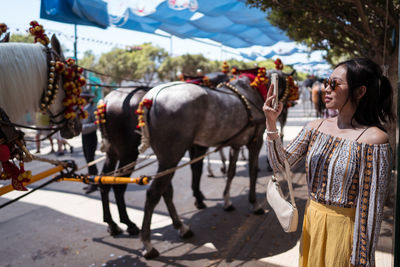 Asian female tourist with fan shooting mules with decorated bridles on sunlit street during fair in seville, spain