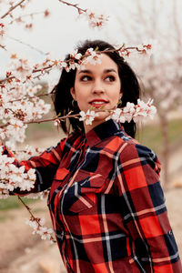 Young woman looking away while standing by cherry tree