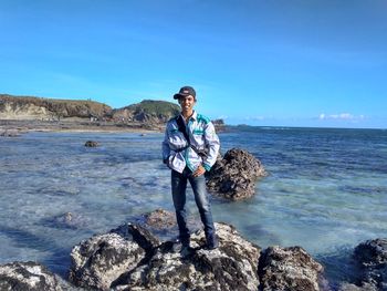 Portrait of young man standing on rock by sea against blue sky