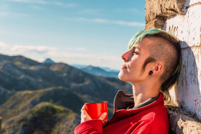 Side view of androgynous traveler standing in highlands on sunny day and enjoying hot drink during trip in el mazuco with eyes closed