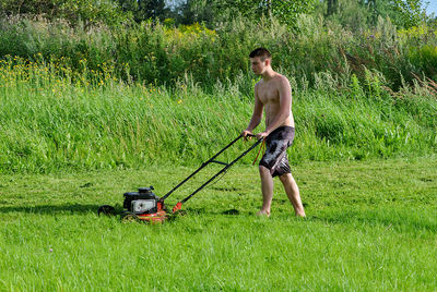 Shirtless young man mowing grassy field