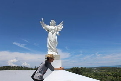 Side view of man wearing hat standing by statue against sky