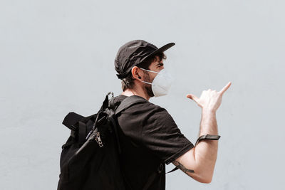 Back view of unrecognizable trendy man with backpack in respiratory mask demonstrating shaka gesture near wall while looking away during coronavirus pandemic