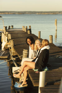 Cheerful young female friends enjoying summer while sitting on jetty at harbor against sky