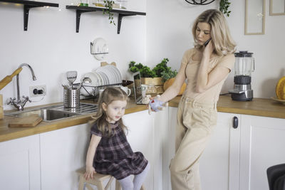 Mother talking on phone while daughter sitting stool in kitchen