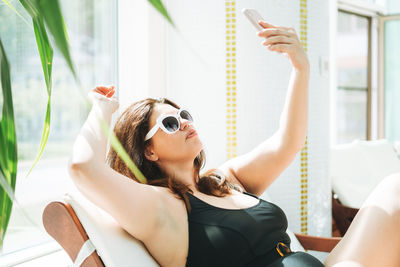 Stylish woman plus size in black swimsuit and sunglasses taking selfie on mobile on lounger in spa