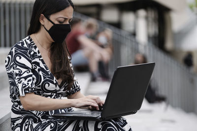 Woman wearing mask working on laptop sitting outdoors