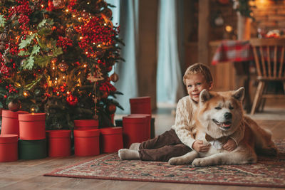 Candid authentic happy little boy in knitted beige sweater hugs dog with bow tie at home on xmas