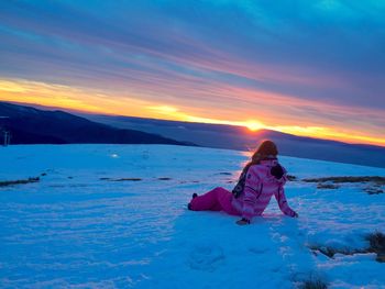 Girl sitting on snow against sky during sunset