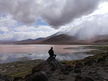 Rear view of man sitting on rock at laguna colorada against cloudy sky